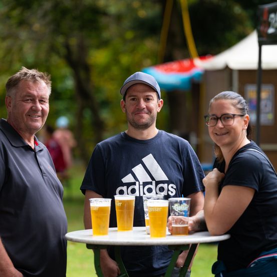 Drei Menschen lächeln vor einem Tisch mit Biergläsern. Ein Mann und eine Frau stehen auf beiden Seiten eines Mannes mit einer Kappe. Ein Zelt und Bäume im Hintergrund.