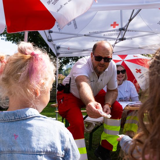 Ein Mann in reflektierender Kleidung reicht einem Mädchen mit rosa Haaren ein Papier unter einem Zelt. Eine Frau mit Sonnenbrille sitzt in der Nähe.