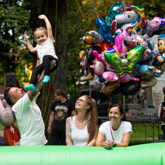 Eine fröhliche Familie spielt auf einem grünen Aufblasbarem bei einer Outdoor-Veranstaltung. Ein Mann hebt ein kleines Mädchen hoch, während zwei Frauen in der Nähe lächeln. Bunte Luftballons und Bäume im Hintergrund.