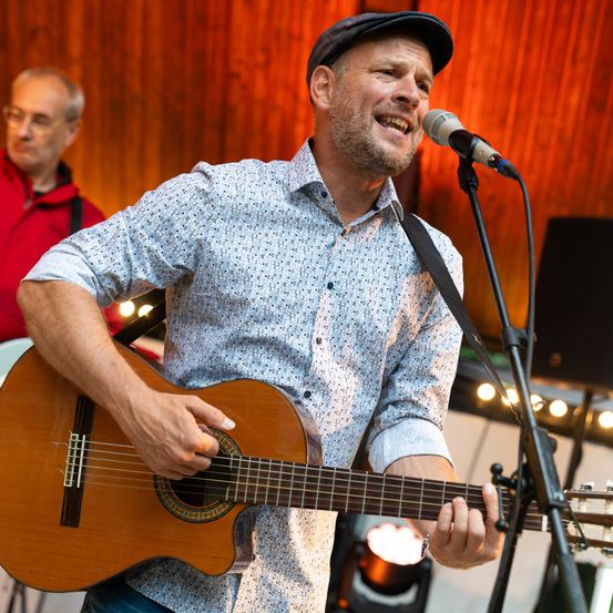 Ein Mann spielt Gitarre und singt in ein Mikrofon auf einer Bühne. Er trägt eine Mütze und ein hellblaues Hemd. Ein Mann in einem roten Jackett steht hinter ihm.