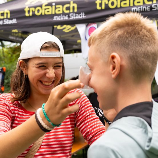 Eine lächelnde Frau in einem rot gestreiften Shirt pustet spielerisch eine Blase in das Gesicht eines jungen Jungen, der amüsiert aussieht. Sie stehen unter einem Vordach mit 'trofaiaich meine Stadt' Beschilderung.