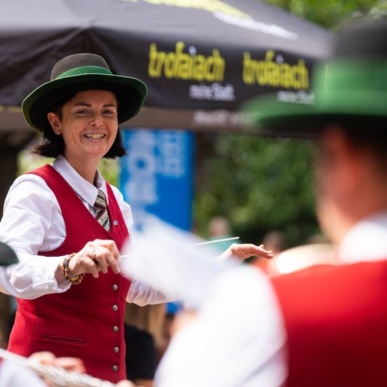 Eine Frau in einem roten Wams und grünem Hut lächelt und spielt mit einem Taktstock bei einer Parade.