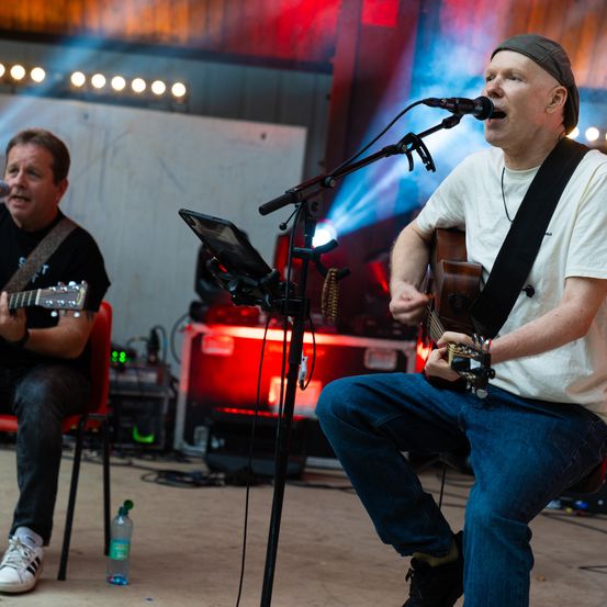Zwei Männer treten auf der Bühne auf, einer singt und spielt Gitarre, der andere spielt Gitarre, mit Scheinwerfern und Ausrüstung im Hintergrund.