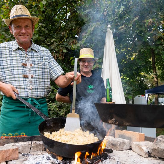 Zwei Männer kochen im Freien mit einem Feuer. Einer wendelt das Essen mit einem Kochlöffel, der andere hält ein Messer. Eine Flasche und ein weißer Schirm sind in der Nähe.