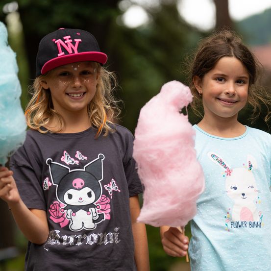 Zwei Mädchen stehen draußen und halten Zuckerwatte. Das Mädchen auf der linken Seite trägt eine schwarze Kappe mit rosa Buchstaben, während das Mädchen auf der rechten Seite eine rosa Zuckerwatte hält.