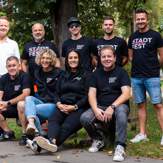 Eine Gruppe Erwachsener in Stadt Fest T-Shirts posiert für ein Foto im Park. Sie sitzen auf einer Bank mit einem Baumstamm dahinter.