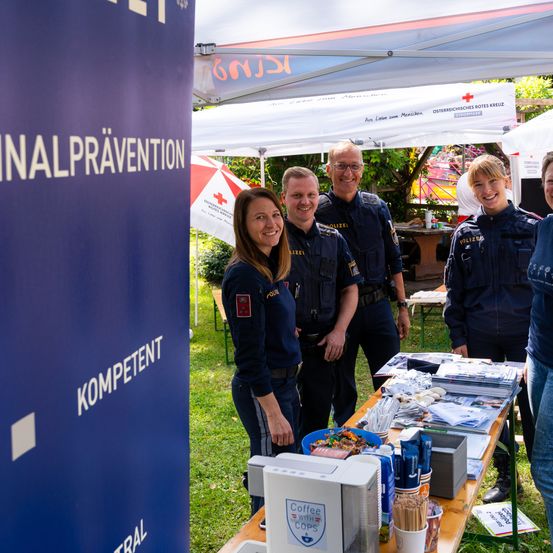 Eine Gruppe von Polizeibeamten steht in einem Stand unter einem Zelt und lächelt für ein Foto. Ein Tisch mit Gegenständen und einer Kaffeemaschine ist sichtbar.