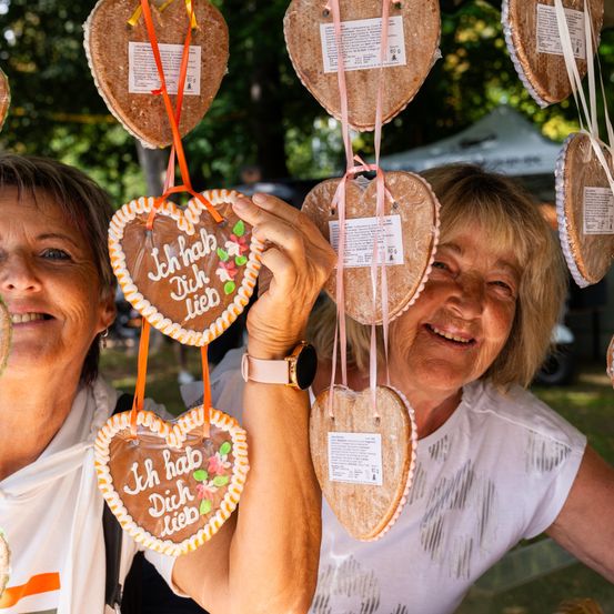 Zwei Frauen lächeln, während sie herzförmige Kekse hochhalten, auf denen 'Ich hab Dich Lieb' steht. Sie sind im Freien in einem Park.