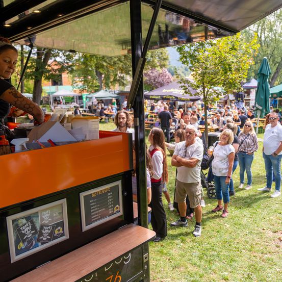 Ein Imbisswagen mit einer Speisekarte steht unter einem Vordach in einem Park. Kunden stellen sich an, um Essen zu bestellen. Eine Frau mit einem Tattoo serviert das Essen.