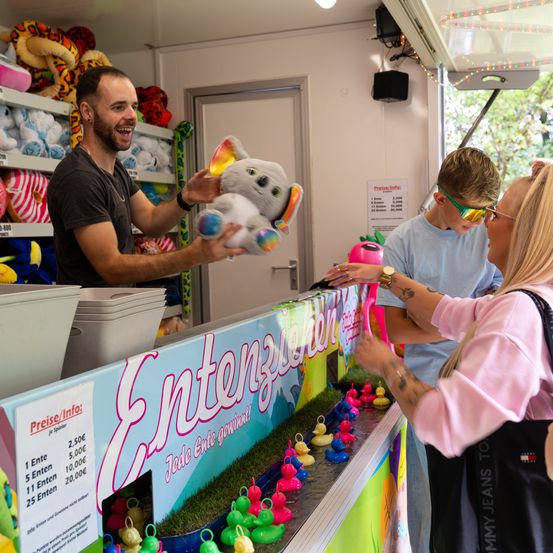 Ein Mann gibt einer Frau und ihrem Sohn ein Kuscheltier in einem Stand. Der Stand hat ein Schild für Entenwerfen. Ein Junge trägt eine Sonnenbrille.