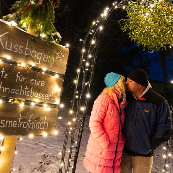 Ein Paar küsst sich unter einem beleuchteten Holzschild mit Dekorationen in einem verschneiten Bereich. Das Schild steht für 'Kissbogen'.