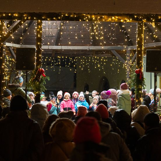 Eine Gruppe von Kindern tritt nachts auf der Bühne auf, umgeben von einem Publikum, unter einem Pavillon, der mit festlichen Lichtern geschmückt ist.