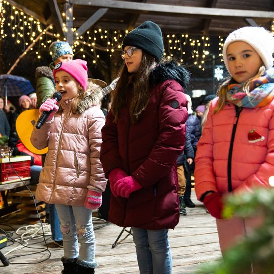 Drei junge Mädchen in Wintermänteln singen in einer festlichen Umgebung mit Lichterketten über ihnen. Eine hält ein Mikrofon, und andere beobachten im Hintergrund.