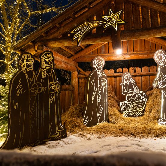 Eine Krippe in einer Holzscheune bei Nacht mit Sternendekoration, Heu und illuminierten Figuren. Die Szene zeigt Maria, Josef, Jesus und drei Weise.