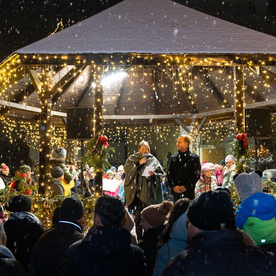 Ein nächtliches Treffen unter einem Pavillon mit Weihnachtsbeleuchtung. Eine Frau hält ein Mikrofon und ein Papier, während ein Mann hinter ihr steht. Es schneit und viele Menschen sind versammelt.