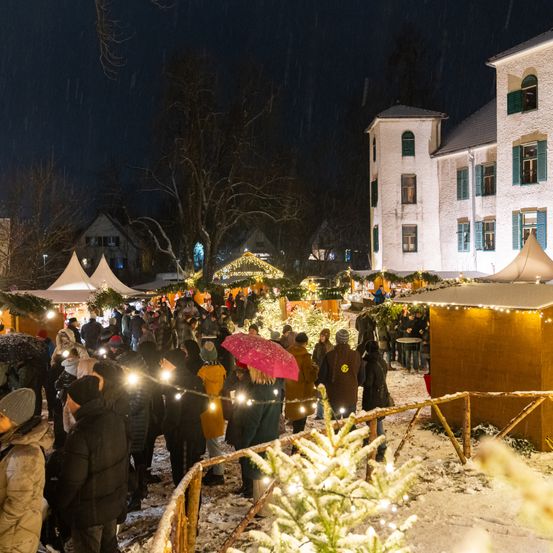 Ein überfüllter Weihnachtsmarkt im Freien mit Menschen, die Lichter halten, beleuchteten Zelten und einem Gebäude im Hintergrund bei verschneiten Wetterbedingungen.