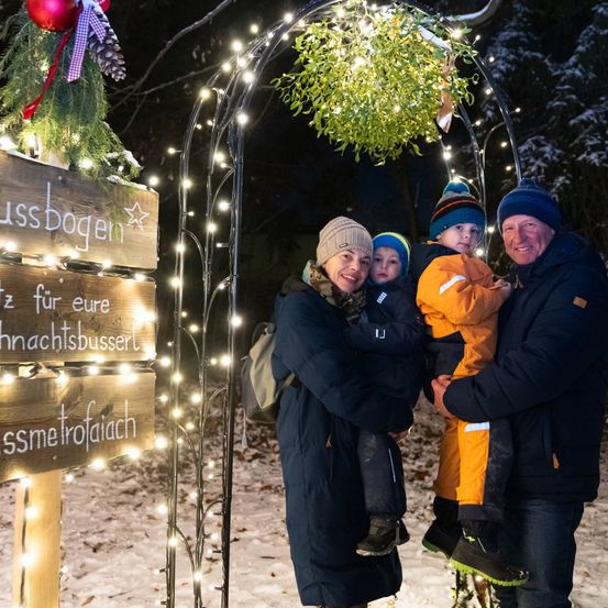 Eine Familie steht im Winter draußen, lächelt und ist von Lichtern und Dekorationen umgeben, darunter ein Schild mit der Aufschrift 'ussboggen' und 'z fur eure nachtsbusser'.