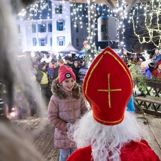 Ein Mädchen in Winterkleidung steht vor einer Figur, die als Sankt Nikolaus gekleidet ist. Sie befinden sich in einer festlichen Umgebung im Freien mit Lichtern und einer Menschenmenge.