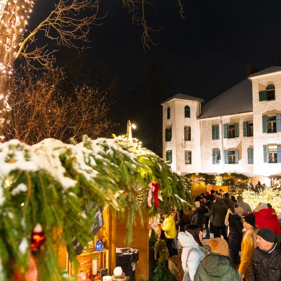 Bei Nacht laufen mehrere Menschen in Winterkleidung auf einem Freigelände. Es gibt Weihnachtsbäume und Girlanden. Ein weißes Gebäude befindet sich hinter ihnen.