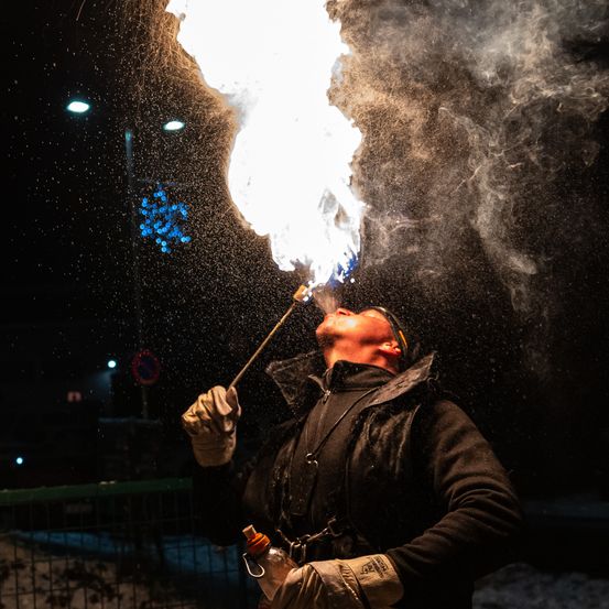 Ein Mann bläst nachts Feuer aus einer Röhre. Er trägt einen schwarzen Mantel und Handschuhe.