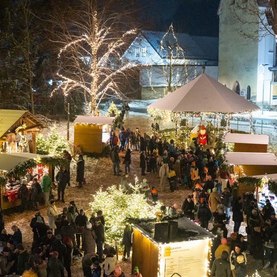 Eine nächtliche Szene eines geschäftigen Weihnachtsmarktes. Der Markt ist mit festlichen Lichtern, Bäumen und zahlreichen Ständen gefüllt. Menschen gehen herum und genießen das warme Leuchten der Lichter und die festliche Atmosphäre.