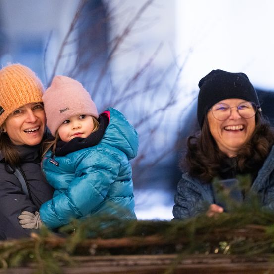 Drei Frauen und ein junges Mädchen stehen lächelnd zusammen, warm gekleidet, mit einem verschneiten Hintergrund.