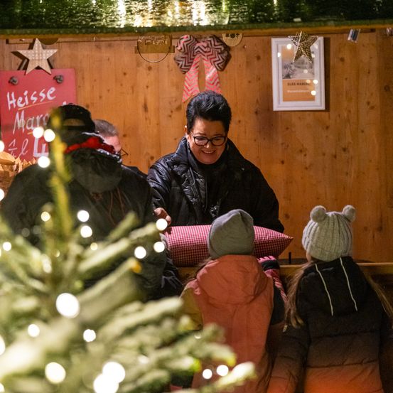 Eine lächelnde Frau steht an einem Stand, möglicherweise verkauft sie heiße Schokolade, mit zwei Kindern und zwei Erwachsenen vor ihr. Der Stand hat Holzwände und einen Weihnachtsbaum mit Lichtern.