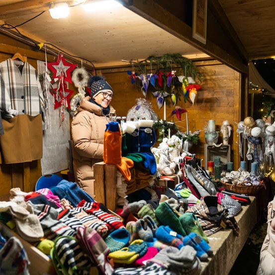 Eine Frau mit Brille und Mütze steht bei Nacht in einem Geschäft. Auf dem Tisch sind verschiedene Artikel ausgestellt, darunter Socken, Mützen und andere Gegenstände. Mehrere Dekorationen hängen an der Wand.