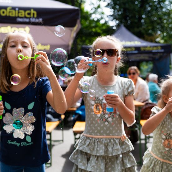 Drei junge Mädchen pusten im Freien Seifenblasen. Das Mädchen auf der linken Seite trägt ein blaues T-Shirt mit Blumendesign. Das mittlere Mädchen trägt ein ärmelloses Oberteil mit Blumenmuster. Das Mädchen auf der rechten Seite hat die Hände an den Wangen. Im Hintergrund sitzen Leute an einem Tisch.