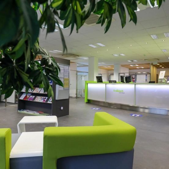 Eine moderne Lobby mit grünen Stühlen und einer Rezeption mit 'troiajiach' darauf. Ein Bücherregal mit Büchern steht links und Monitore sind auf dem Schreibtisch.