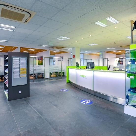 Ein modernes Büroinnenraum mit Rezeptionstresen, Monitoren und einem Bücherregal. Der Boden ist gefliest, und es gibt Abstandsaufkleber auf dem Boden.