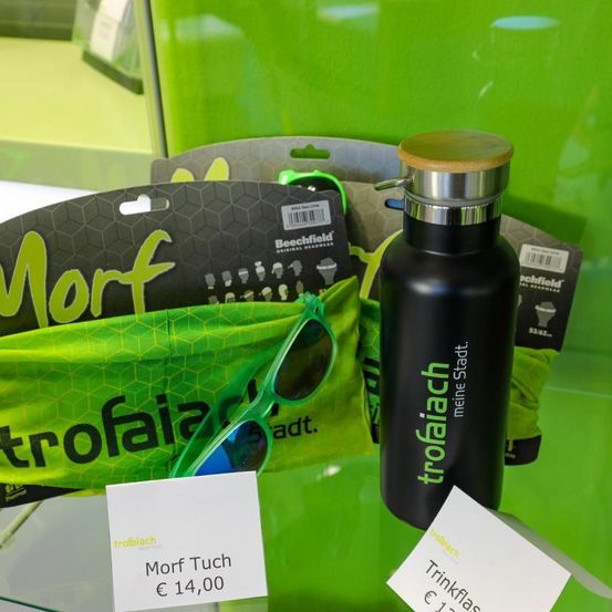 Ein Morf-Handtuch wird zusammen mit einer Tropich-Flasche, einer Sonnenbrille und anderen Gegenständen auf einem Glasregal ausgestellt. Das Handtuch ist grün mit dem Wort Morf in weißen Buchstaben. Die Flasche hat das Wort Tropich in Grün auf einem schwarzen Körper.