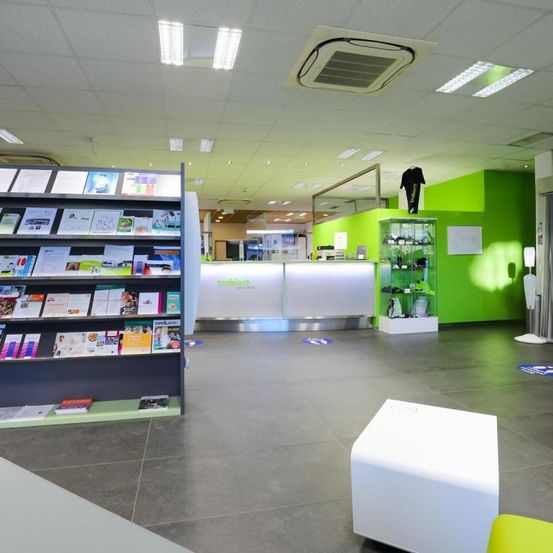 Ein Innenraum eines modernen Büros mit grünen Wänden und weißer Decke. Ein Regal mit Büchern und Zeitschriften ist links. Ein Schreibtisch mit weißer Oberfläche und ein Glasvitrine steht in der Mitte. Ein weißer kubischer Hocker ist rechts.