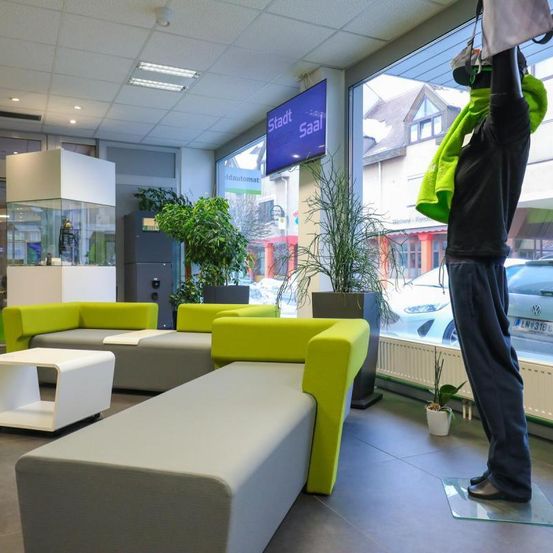 Ein Büroinnenraum mit einer Person in Virtual-Reality-Ausrüstung. Grüne Sofas, weiße Tische und Pflanzen schmücken den Raum. Ein Fernseher und ein Monitor sind an der Wand montiert. Draußen sind Autos und Häuser durch ein großes Fenster sichtbar.