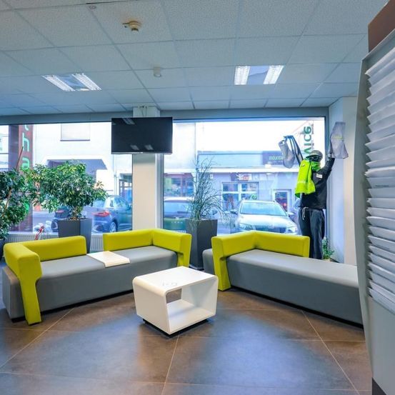 Ein modernes Büro-Interieur mit zwei gelben und grauen Sofas, einem weißen Tisch und großen Fenstern. Eine Person steht am Fenster.