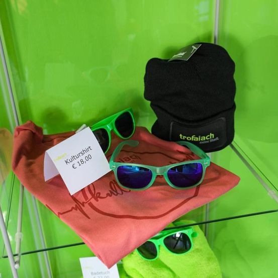 Ein roter Stoff, eine schwarze Mütze und drei Paar Sonnenbrillen auf einem Glasregal vor grünem Hintergrund. Der rote Stoff hat einen weißen Aufkleber mit der Aufschrift 'Kulturshirt 18,00'. Die Mütze hat 'trofaiaich' darauf geschrieben. Die Sonnenbrillen sind grün mit blauen Gläsern.