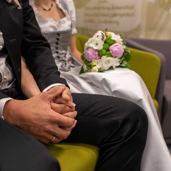 Ein Paar in formeller Kleidung sitzt auf einem Stuhl, hält Händchen und neben ihnen steht ein Blumenarrangement. Der Mann trägt einen schwarzen Anzug und die Frau ein weißes Kleid.