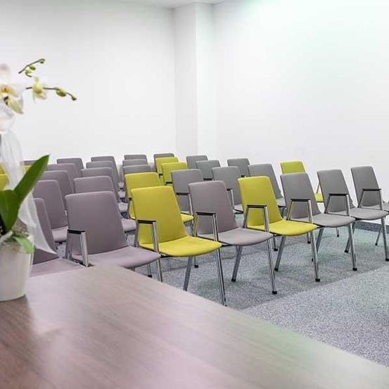 Ein leerer Konferenzraum mit grauen und gelben Stühlen in Reihen, einem Holztisch mit einer weißen Vase mit weißen Blumen und weißen Wänden.