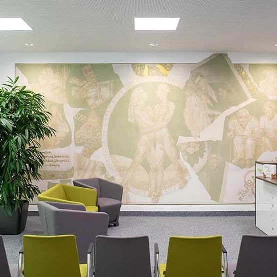 Ein Raum mit einer großen Wandmalerei, mehreren Stühlen, einem Tisch und einer Topfpflanze. Die Wandmalerei zeigt verschiedene Szenen.