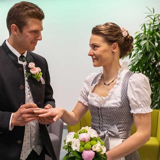 Ein Mann und eine Frau in traditioneller Kleidung tauschen einen Ring aus. Sie halten Händchen und lächeln sich an. Die Frau hält einen Blumenstrauß in der Hand.
