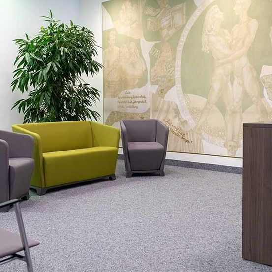 Ein leerer Raum mit grauen Sesseln und einem gelben Sofa. Eine Pflanze steht in der Ecke. Eine große Wandmalerei ist an der Wand. Eine Blume steht in einer Vase auf dem Schreibtisch.