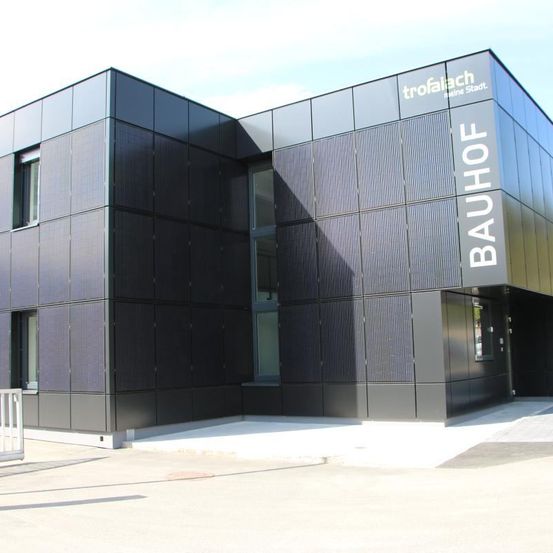 Ein modernes Gebäude mit Solarpaneelen an der Außenseite, mit einem Schild, das 'BAUHOF' liest. Das Gebäude besteht aus schwarzen Paneelen und hat Glasfenster.