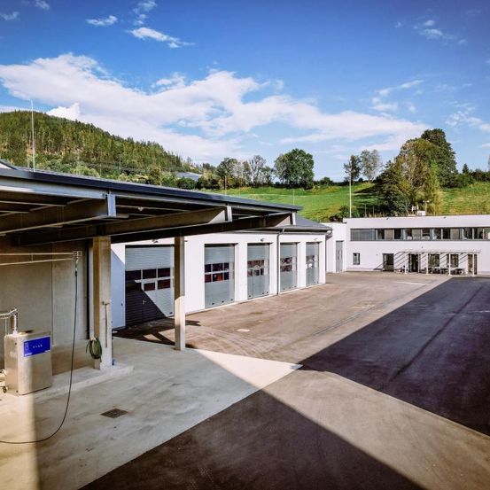 Ein Freigelände mit einem Parkplatz unter einem Vordach, mit mehreren Garagen und einem Gebäude in der Ferne. Der Himmel ist blau mit Wolken.