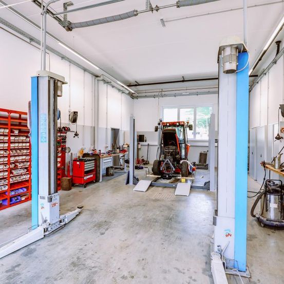 Eine große Garage mit einem roten Traktor unter einer blauen Hebebühne. Der Raum ist gut organisiert mit Werkzeugen, Regalen und Aufbewahrungsboxen. Die Decke hat Metallträger und Leuchtstofflampen.