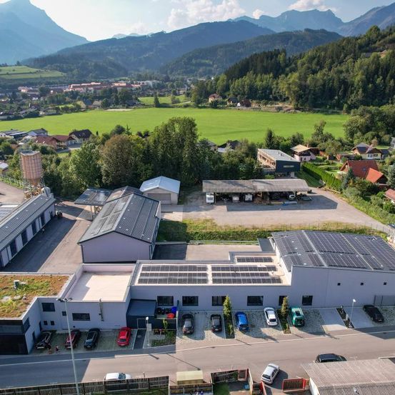 Ein Luftbild einer Fabrik mit Solarmodulen auf dem Dach. Es ist von einem Parkplatz mit mehreren Autos umgeben. In der Nähe befinden sich Häuser und ein Feld. Berge sind in der Ferne.