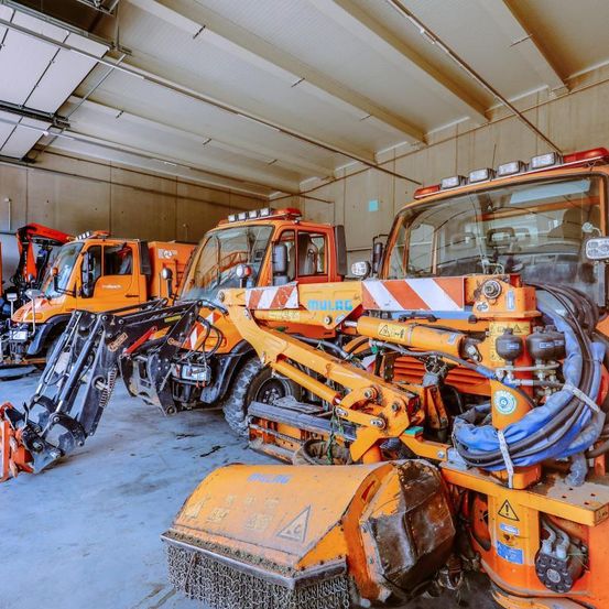 Eine Garage mit mehreren orangefarbenen Schneepflügen geparkt. Einer hat eine gelbe Front. Ein anderer hat ein gelb-schwarz-gelbes Design an der Seite.
