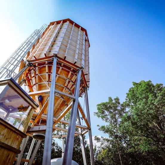 Ein hölzerner Wasserturm auf Metallbeinen hat ein Glasfenster auf einer Seite. Er wird von Bäumen unter einem klaren blauen Himmel umgeben.