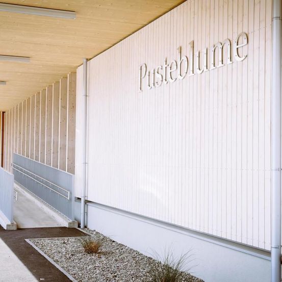 Eine weiße Wand mit dem Wort 'Pisteblume' in großen weißen Buchstaben. Darunter befindet sich ein Kiesbereich mit einer Pflanze. Die Wand hat vertikale Holzpaneele.