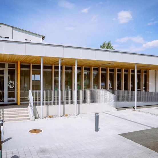 Ein modernes Gebäude mit weißer Außenseite, Holzverkleidung und großen Glasfenstern ist an einem sonnigen Tag zu sehen. Es hat einen geräumigen Eingang mit Treppen, einem gepflasterten Gehweg und einem klaren blauen Himmel.