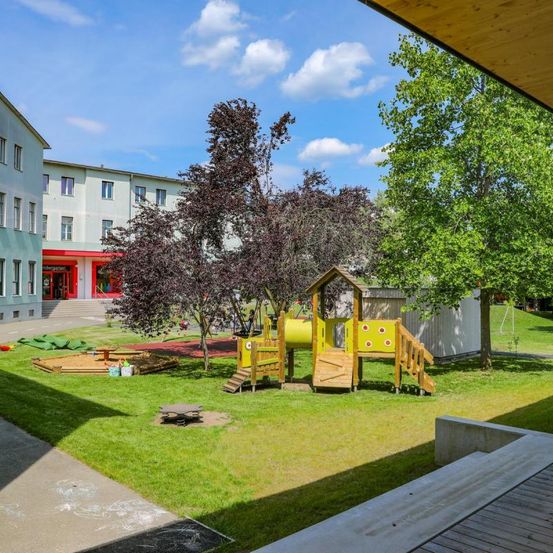 Ein Spielplatz mit einer gelben Rutsche und Klettergerüst steht in einer Rasenfläche, mit einem Gebäude und Bäumen im Hintergrund.